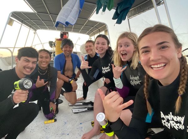 Group in dive suits smiles at the camera while kneeling on boat.