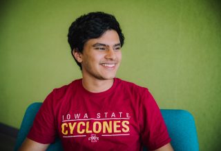 Student in red Iowa State Cyclones t-shirt sits in a blue chair, with a green wall in the background.