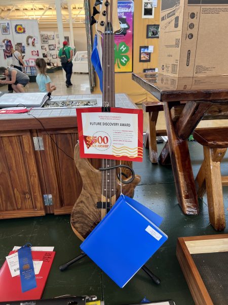 A built bass guitar is displayed in the 4-H exhibits building at the Iowa State Fair. Attached to the guitar is an LAS Future Discovery Award certificate and a blue binder.