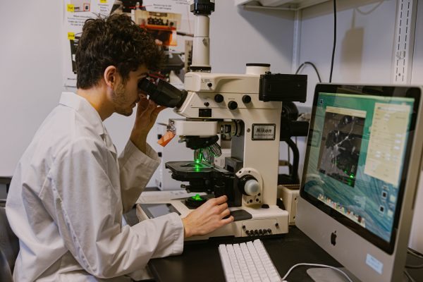 Student in white lab coat looks into a laboratory microscope. Imagery is displayed on a computer screen next to the student.